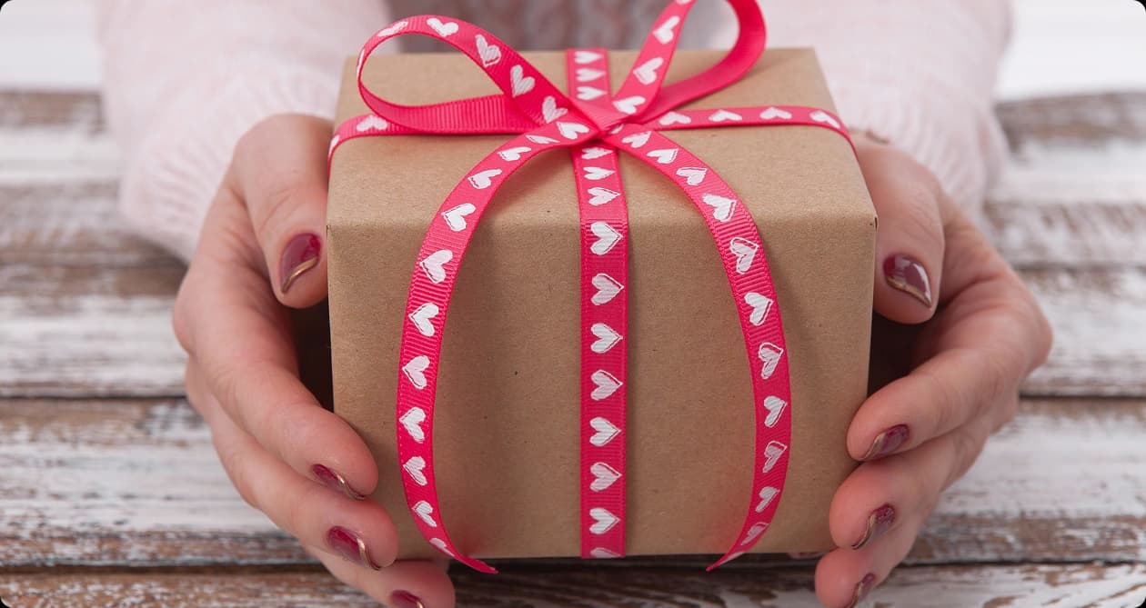 Hands presenting a wrapped gift box with a heart-patterned ribbon